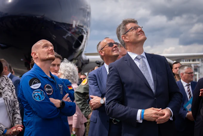 Auf dem Bild sieht man Bundesverkehrsminister Patrick Schnieder zusammen mit Bundeskanzler Friedrich Merz und einem Mitarbeiter der European Space Agency, wie sie auf der ILA 2026 in Berlin in den Himmel nach rechts schauen und eine Flugschau beobachten.