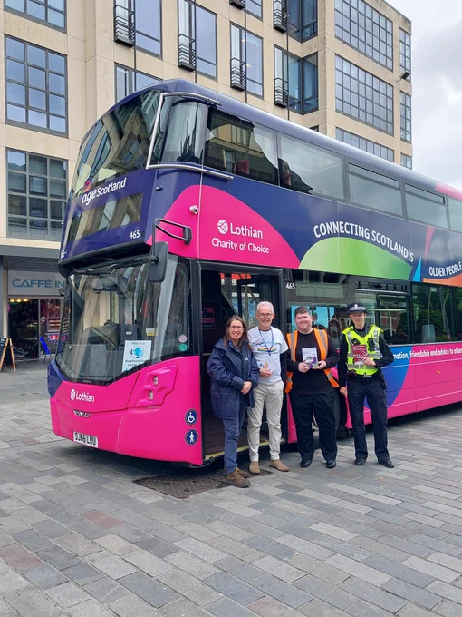 A group of five people, including a police officer, stand beside a pink and blue Lothian double-decker bus.