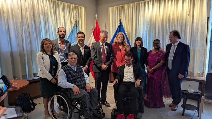 Group photo at the Austrian Permanent Mission to the United Nations during the side event, “Startups and Emerging Technologies as Drivers of Disability Human Rights? Looking Ahead to the CRPD’s Third Decade.” Eleven attendees pose together in a meeting room, with United Nations, Austrian, and other national flags displayed behind them. Two participants are seated in mobility devices at the front of the group, while others stand behind them 