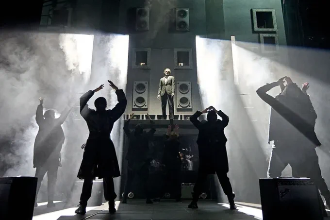 On a dark, smokey stage, silhouetted figures hold their hands up toward a figure standing high on a platform