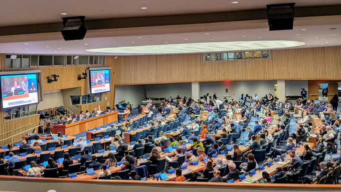Wide view of a United Nations conference chamber during a Conference of States Parties (COSP) session focused on disability rights. Hundreds of delegates, advocates, government representatives, and observers are seated throughout the auditorium, facing a panel of speakers at the front. Large screens display the live discussion with captions and sign language interpretation. The room is equipped with individual desks, microphones, and accessibility supports, reflecting the international gathering’s focus on disability inclusion and policy. 