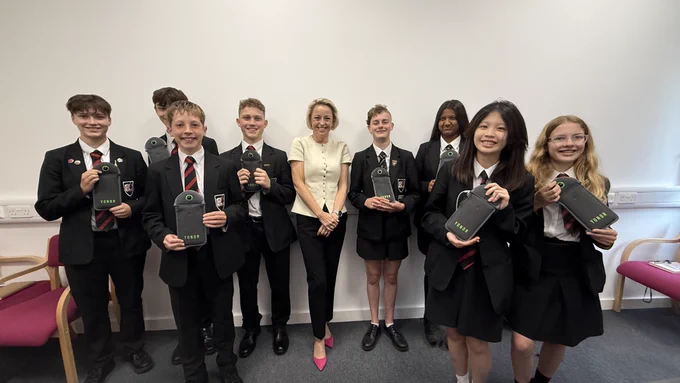 Image shows the Police and Crime Commissioner Lisa Townsend who is a blonde short-haired woman wearing a lemon coloured blouse and black slim cropped trousers standing in a small meeting room amongst 8 male and female year 9 students wearing the The Winston Churchill School uniform.  This uniform is black trousers or skirt, white shirt, black blazer and red and black striped tie. Each student in the picture is holding up a small grey fabric mobile phone pouch in front of them. 