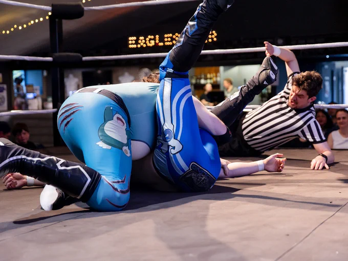 Two competitors at the Super Effective event in Bedford, tightly tangled in a wrestling match, with the referee behind them on the floor, about to end the match and announce the victor.
