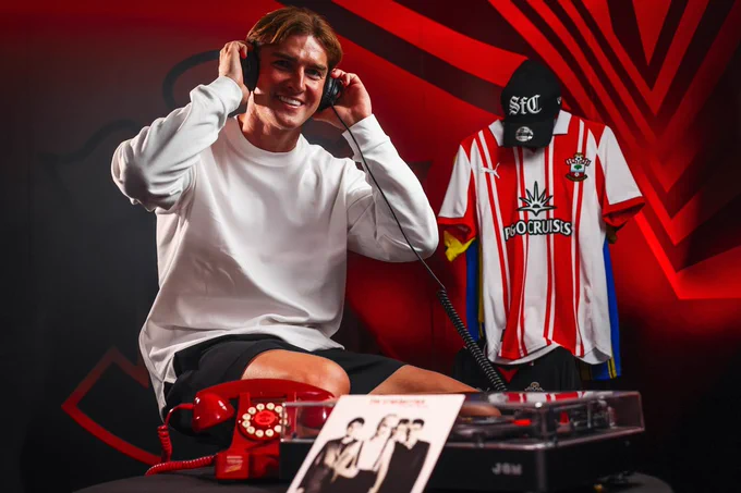 James Bree wearing headphones whilst smiling towards the camera. There is a Saints telephone and a record player with the vinyl of The Cranberries in front of him.