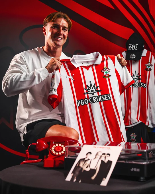 James Bree holds up a Southampton home shirt whilst smiling towards the camera. There is a Saints telephone and a record player with the vinyl of The Cranberries in front of him.