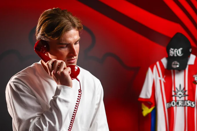 James Bree holding the telephone to his ear. There is a Saints shirt hung up behind him along with a cap that says 'SFC' on it.