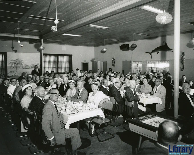 Large group of formally dressed people seated at long tables in a decorated banquet hall with wood-paneled ceiling and mounted fish on the walls.
