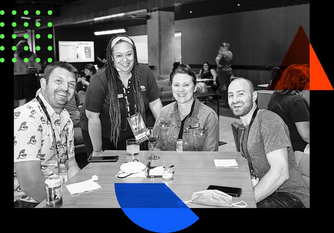 Four people are seen at a table smiling. Three people are sitting, one standing.