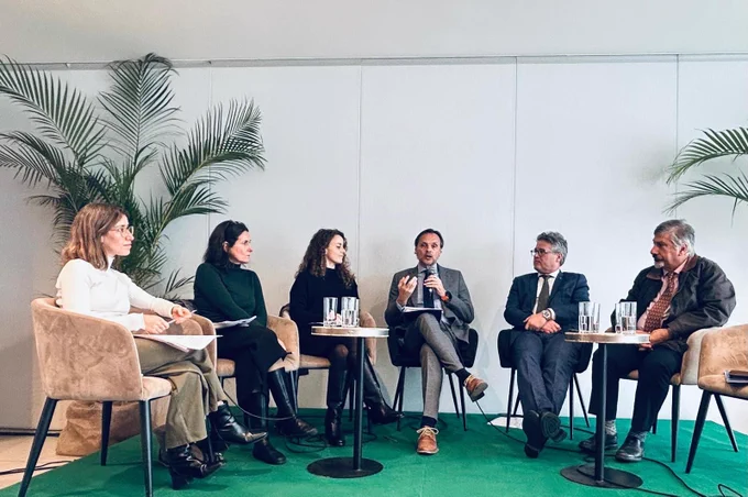 Panel de seis especialistas durante un conversatorio sobre cooperación internacional y gestión del agua. Las personas expositoras están sentadas en semicírculo sobre un escenario, con micrófonos y mesas auxiliares. Al fondo se observan plantas decorativas y un espacio de exposición.