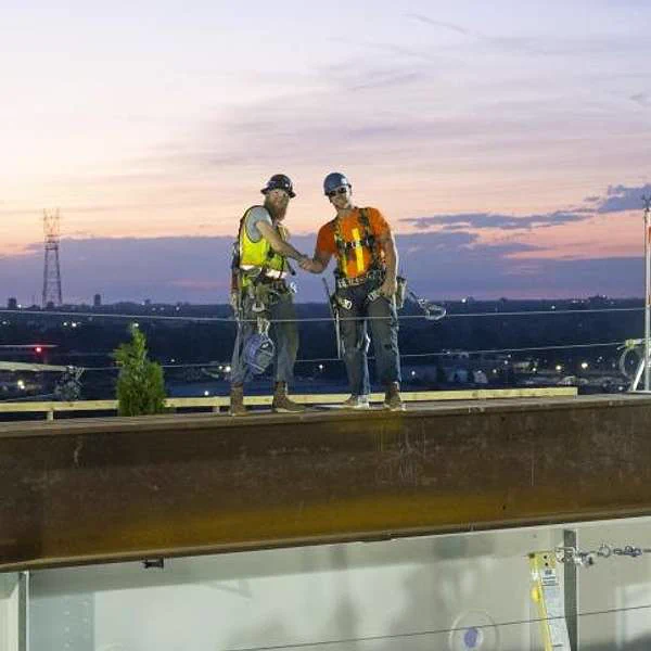 Two men in safety gear shake hands on a bridge beam.
