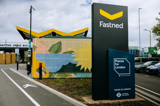 A large black monolith totem features the prominent yellow logo for Fastned alongside branding for Places for London and Transport for London, positioned next to a colourful mural and a distinctively shaped yellow canopy at an electric vehicle charging station near Hatton Cross.