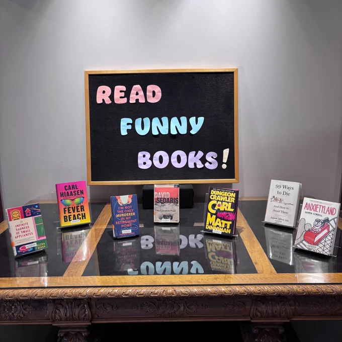 A book display featuring 6 titles: The Infinite Sadness of Small Appliances by Glenn Dixon, Fever Beach by Carl Hiassen, I’m Not the Only Murderer in my Retirement Home by Fergus Craig, The Land and Its People by David Sedaris, Dungeon Crawler Carl by Matt Dinniman, 99 Ways to Die by Ashely Alker, and Anxiety land by Gemma Correll. Signage text: “Read Funny Books!”