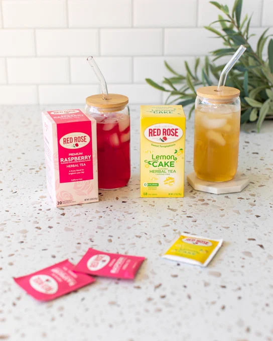 Two glass jars with iced herbal teas, one red rose raspberry and one red rose lemon cake, each with a bamboo lid and glass straw on a terrazzo countertop.