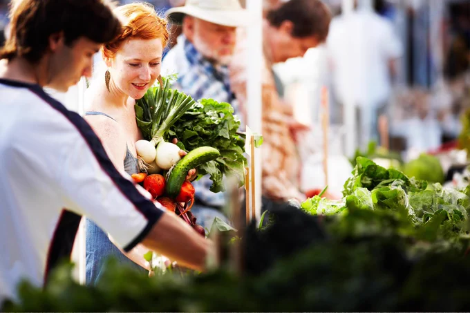 A person holding a bunch of fresh produce while looking at a table full of vegetables with another person. 