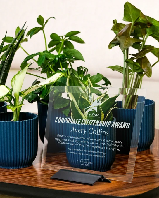 Glass Plaque Engraved With Award Message For Corporate Citizenship Standing On Wood Desk With Plants Via Black Deco Base