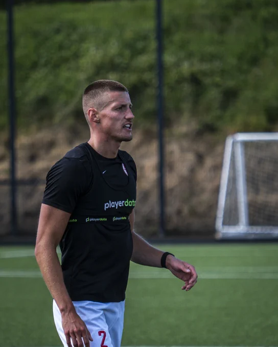 A soccer player in athletic gear is on the field. The player is wearing a black training vest with 'playerdata' visible. A goalpost is in the background.
