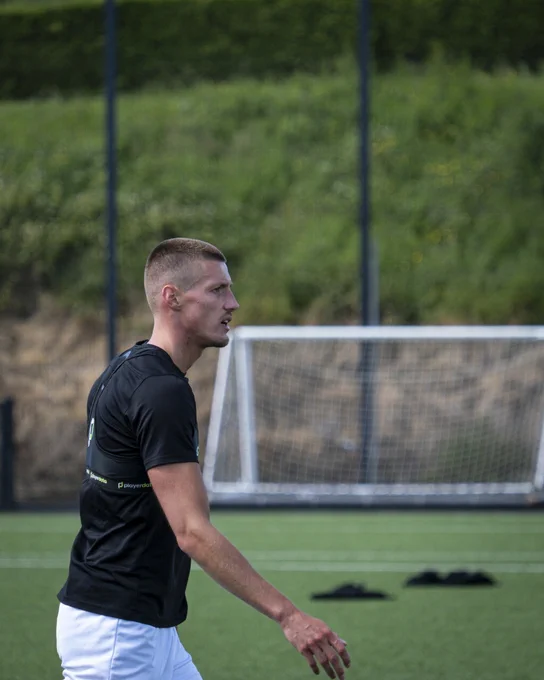 A focused soccer player in a black shirt and white shorts on a green field. The goalpost is visible in the background.