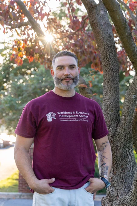 Man in a maroon shirt that reads “Workforce & Economic Development Center” with tattoos stands outside, beside a tree.