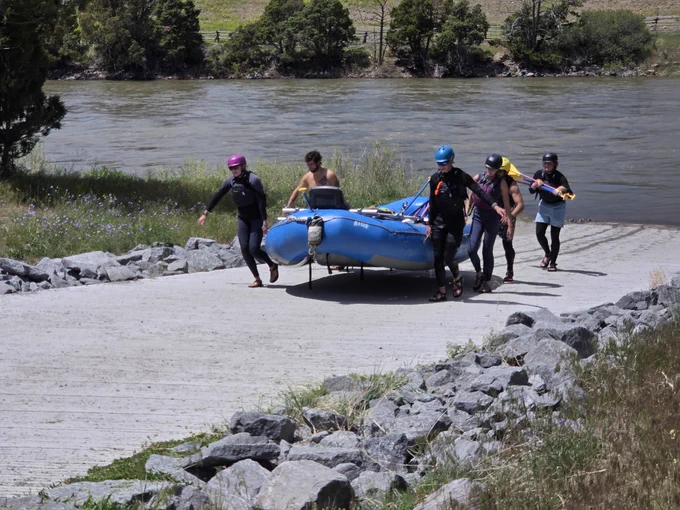 Group of five people carrying a blue inflatable raft toward a river on a rocky path surrounded by greenery.