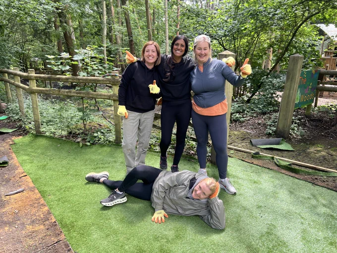 Three people standing on artificial grass in a wooded area with one person lying down in front, all dressed in casual outdoor clothing.