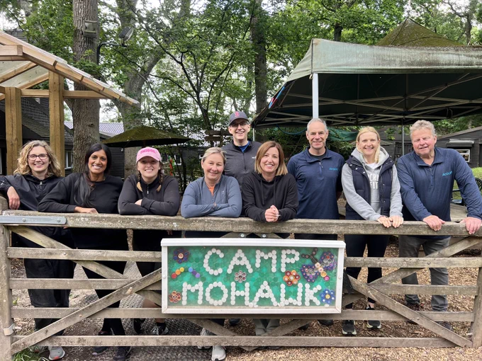 Group of people standing behind a wooden gate with a colorful 'Camp Mohawk' sign in a forested outdoor setting.