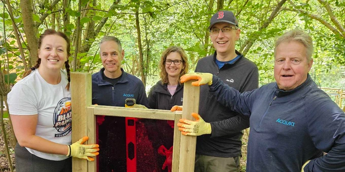 Group of five people standing outdoors around a wooden-framed red panel in a green wooded area.