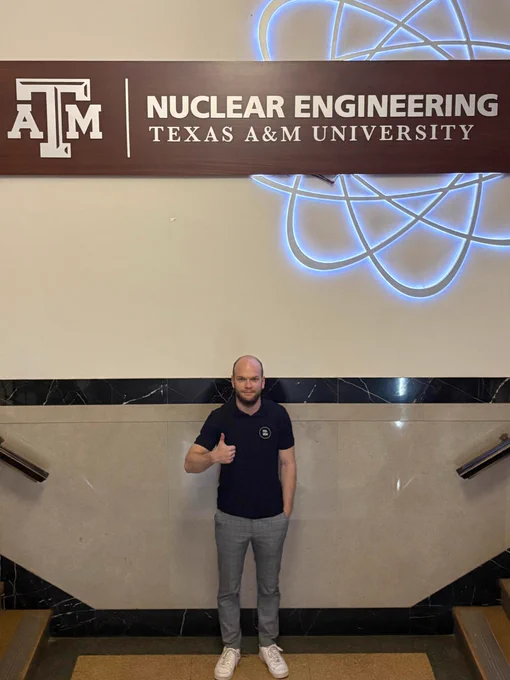 A person stands indoors giving a thumbs-up in front of a sign that reads "Nuclear Engineering, Texas A&M University," with the Texas A&M logo.