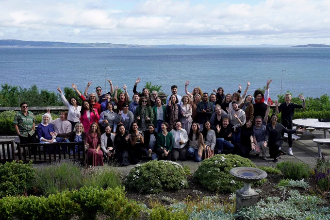 A group imagine of delegates on the Arts Health Research Intensive 2026. Behind the group of delegates is a view of the sea.