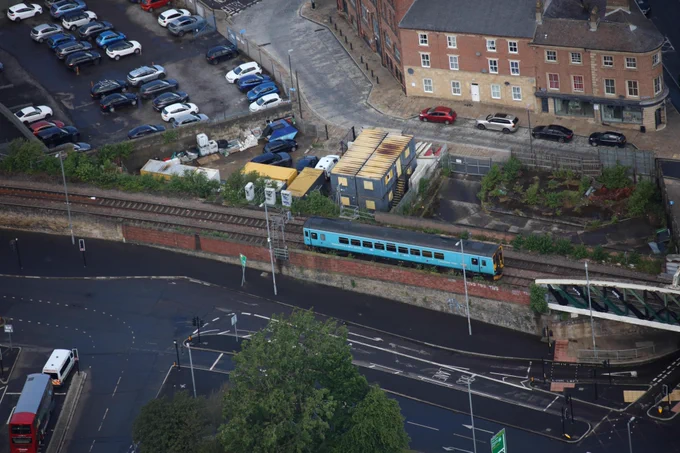 Aerial view of a blue train traveling on a railway. Surrounding the train are parking lots with several parked cars, roads with visible traffic markings.
