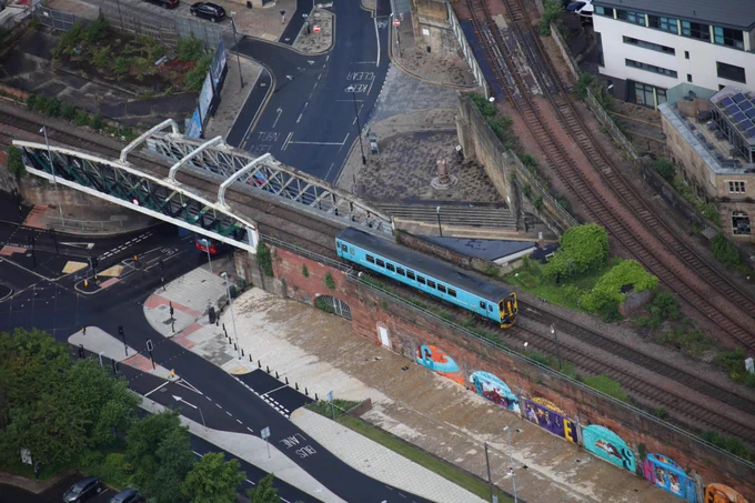 A blue train travels on a railway bridge over a city street, surrounded by green foliage and urban structures. There are colorful murals on the wall beneath the bridge. 