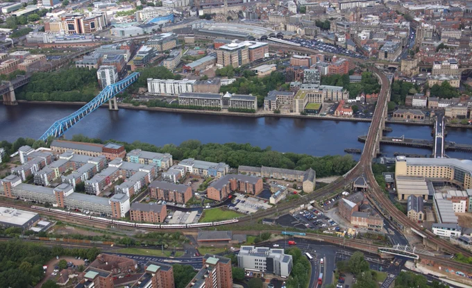 Aerial view of Newcastle upon Tyne, featuring the Tyne Bridge crossing the River Tyne. The cityscape includes a mix of residential and commercial buildings, roads, and railway tracks. 