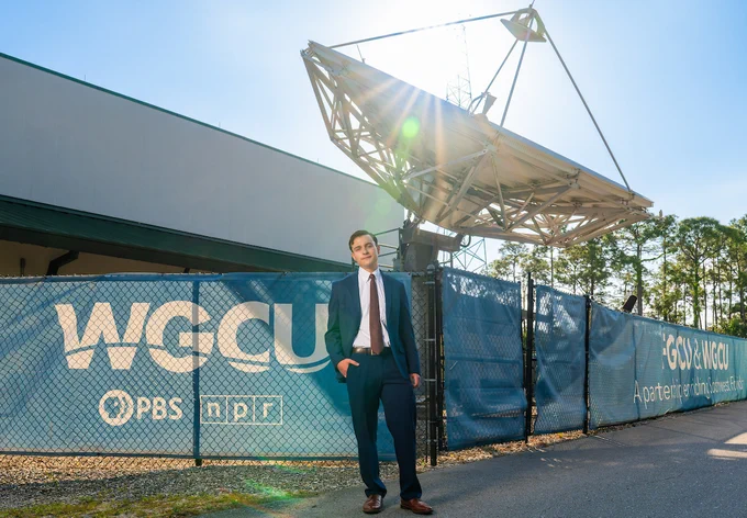 a photo of a student standing outside of a radio station on a sunny day