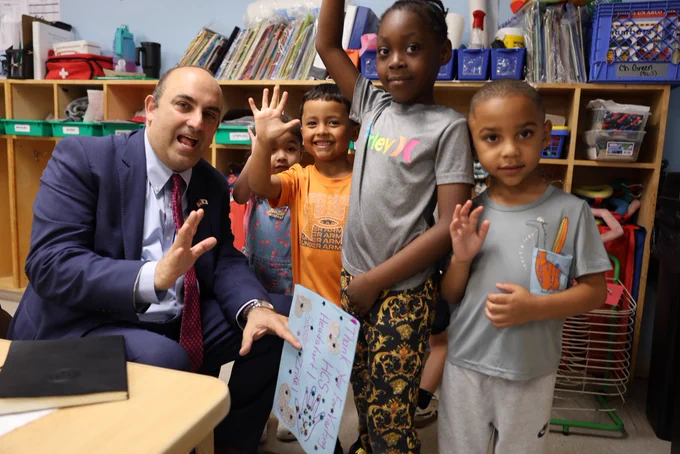 Massachusetts Education Secretary Steve Zrike kneels in a classroom at HCS Head Start in Holyoke, smiling and waving alongside a group of young students. Several children wave back and smile at the camera while standing in front of bookshelves filled with classroom materials, highlighting a warm and engaging early learning environment.