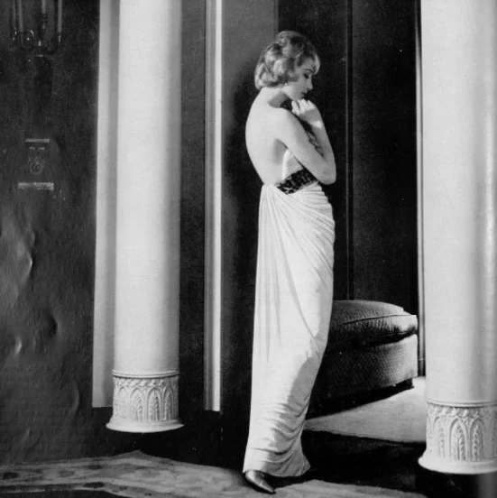 A woman stands thoughtfully by a doorway in an elegant, floor-length gown, with a low back and dark patterned band, amidst classical columns in a softly lit room.