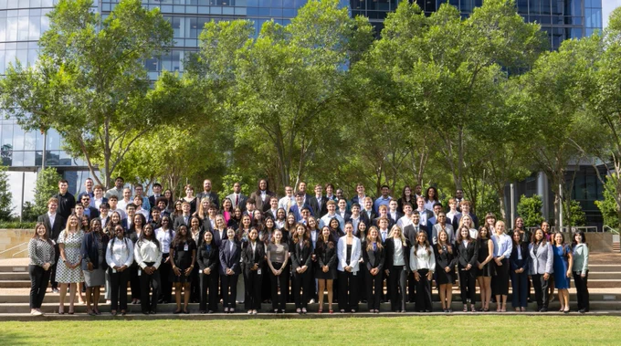 A large group of people is posed together outdoors on steps, with a backdrop of trees and a modern glass building.