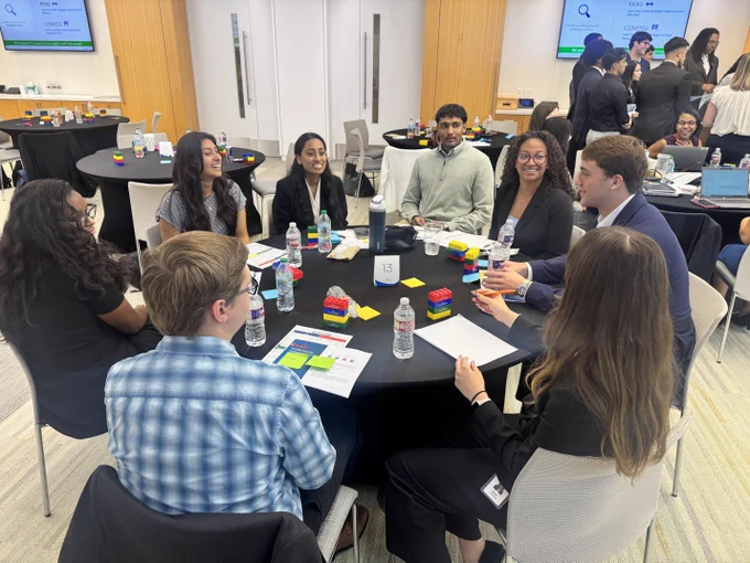 A group of people sitting around a table engaging in discussion. The table has papers, water bottles, and LEGO bricks. In the background, a presentation is visible on a screen.