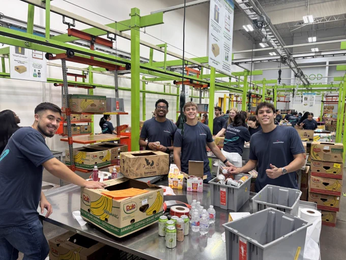 A group of people are packing food items in a warehouse-style setting with shelves and boxes. Various food items and bottled drinks are on the table. The environment appears busy with several people working together.