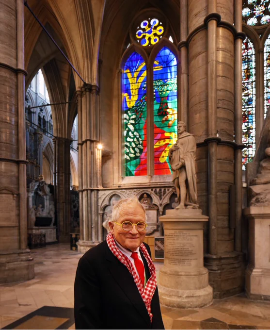 Artist David Hockney - wearing a dark coat, white shirt, red tie, and a red and white scarf - smiling as he stands in front of The Queen's window, the brightly-coloured stained glass window he designed in Westminster Abbey.