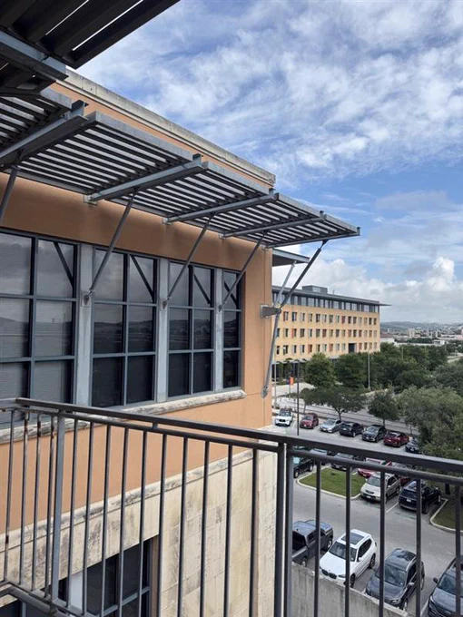 View from a balcony overlooking a parking lot with cars and a multi-story office building under a partly cloudy sky.