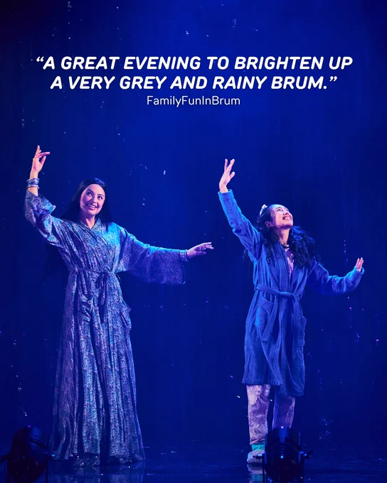 Two performers on stage, dressed in shimmering blue attire, dance gracefully in the rain under a spotlight against a dark background. Above them, a quote reads: “A great evening to brighten up a very grey and rainy Brum.” The quote is attributed to FamilyFunInBrum.