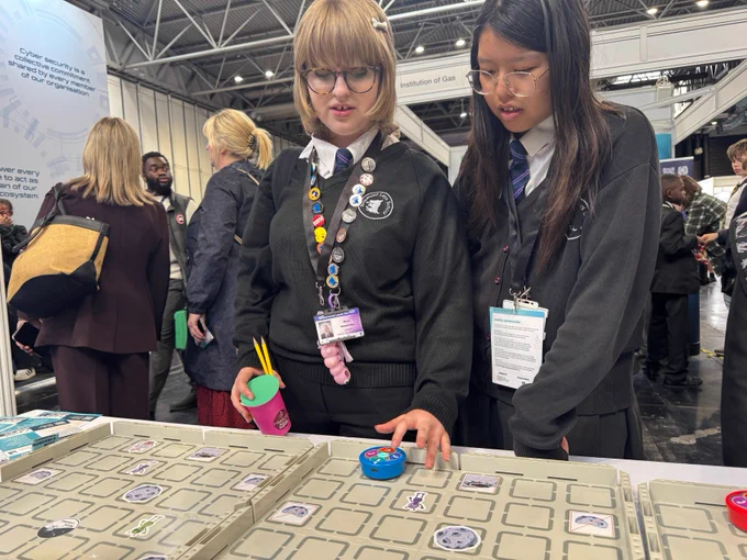 Two individuals in school uniforms interacting with a tabletop educational game. The surface has a grid pattern with various symbols and digital devices. Other attendees and educational posters are visible in the background.