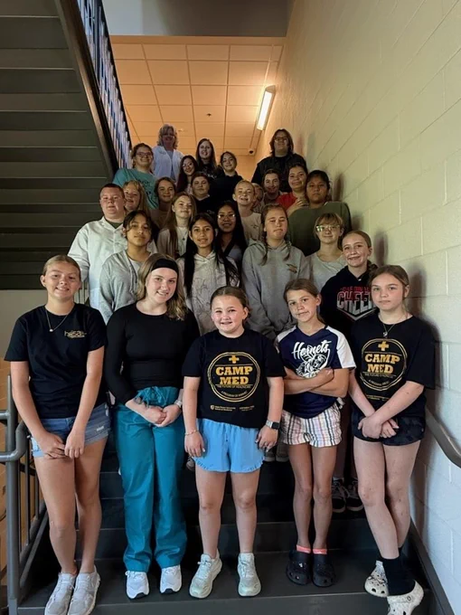 A group of approximately 25 middle school students and two adult leaders pose together on an indoor staircase during Jr. Camp Med. Several students wear Camp Med T-shirts, while others are dressed in casual clothing. The group smiles toward the camera, highlighting a fun and engaging day focused on learning about healthcare careers and future opportunities.