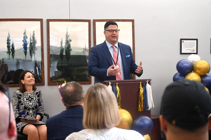 A man speaks at a podium during a Community Justice Support Center ceremony, with attendees seated and listening.