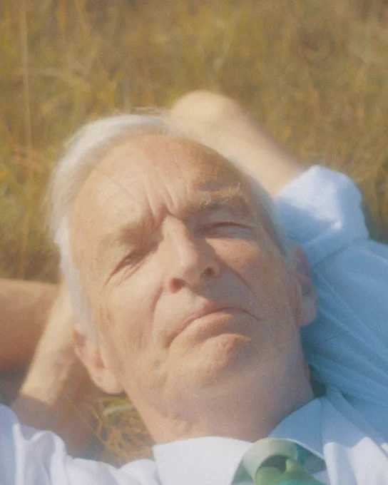 A close up shot of Jon Snow's face and shoulders. He has his eyes closed, and is laying back on some grass. He is wearing a shirt with rolled up sleeves and a green tie.
