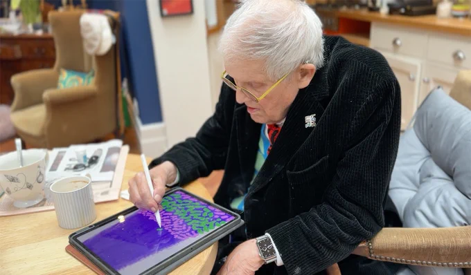 David Hockney sitting at a table, drawing on iPad with Apple Pencil.