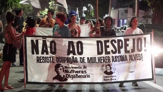 Pessoas seguram em uma rua à noite uma faixa com a frase "Não ao despejo! Resiste Ocupação de Mulheres Rayana Alves!"