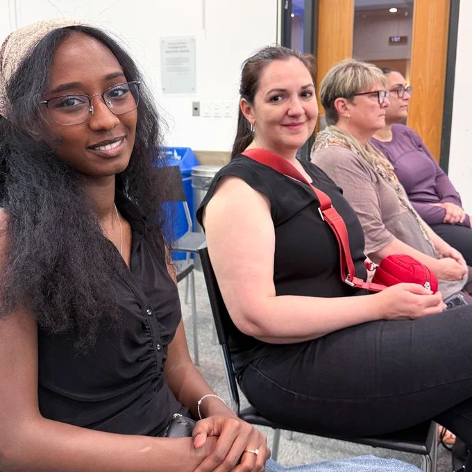 Four attendees seated in a row, one looking toward the camera, others facing forward in a room with chairs.
