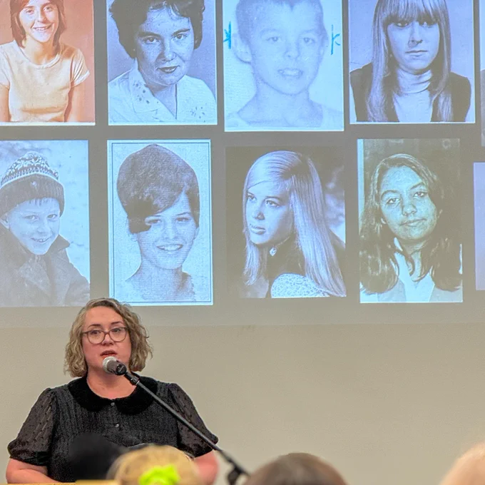 Vanessa Brown speaking at a microphone with a screen of eight blue-tinted portrait photos behind them.
