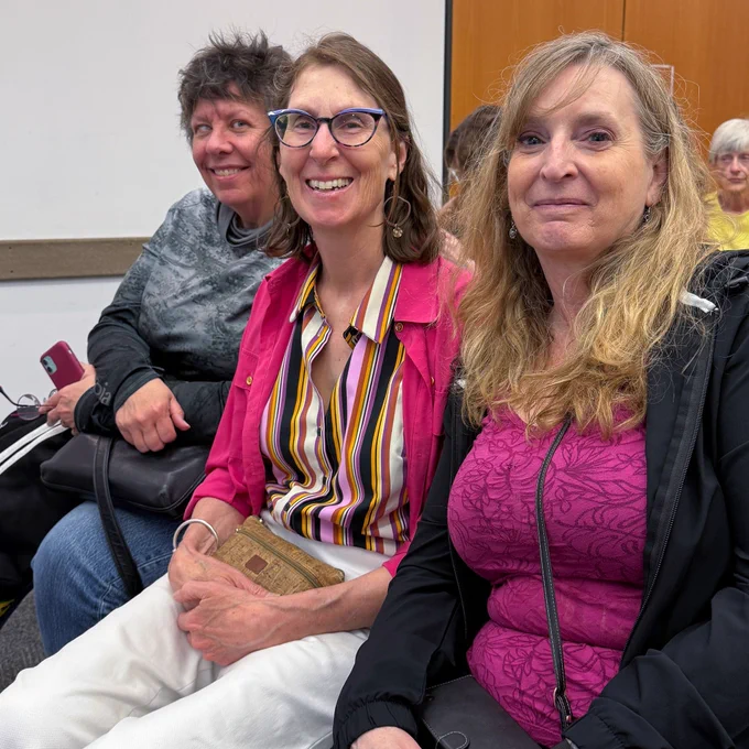 Three attendees seated closely together, smiling at the camera in a meeting room.