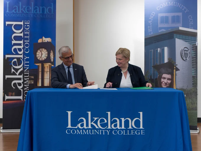 A person and another individual are seated at a table with a blue tablecloth that reads "Lakeland Community College." Behind them are banners featuring the Lakeland Community College logo and images of the campus. They appear to be signing documents.